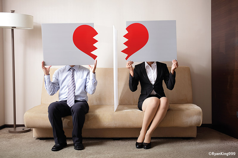 Pärchen sitz auf einer Couch. Die Frau und der Mann halten je ein Schild mit einem gebrochenem Herzen darauf hoch.
